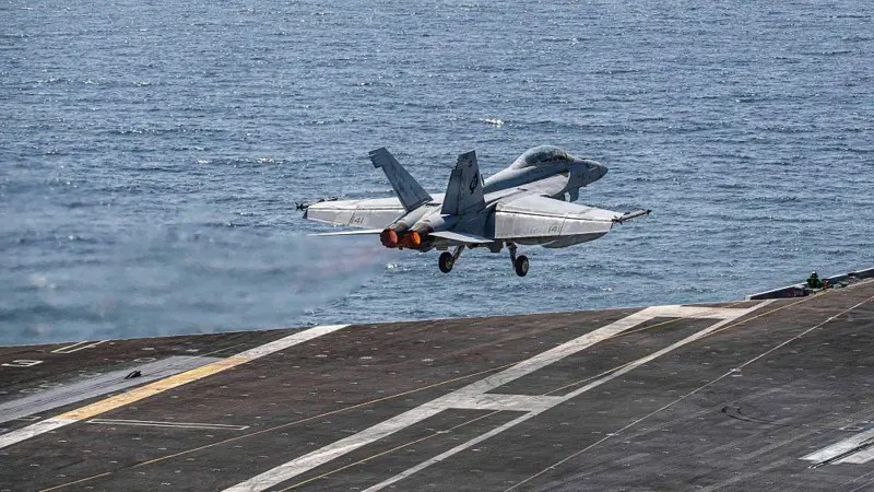 Avião militar de decolagem realizando pouso na pista de um navio no mar, com trem de pouso baixado e rodas tocando o convés, em imagem aérea contra o oceano ao fundo.