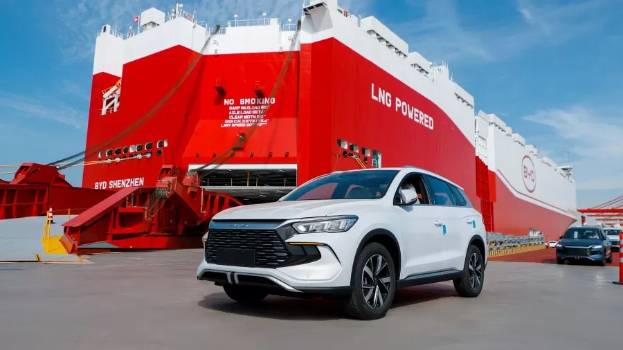Veículo utilitário branco em primeiro plano diante de um navio cargueiro grande com casco vermelho e a placa “LNG POWERED”, em um pátio portuário sob céu azul.