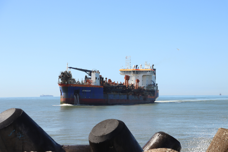 Navio cargueiro atracado ao lado de estruturas portuárias, visto do cais com céu azul e mar calmo ao fundo; a embarcação segue pelo canal.
