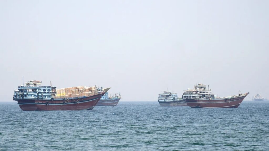 Três navios mercantes e um barco menor navegando no mar em dia nublado, com os cascos grandes visíveis lado a lado no horizonte.