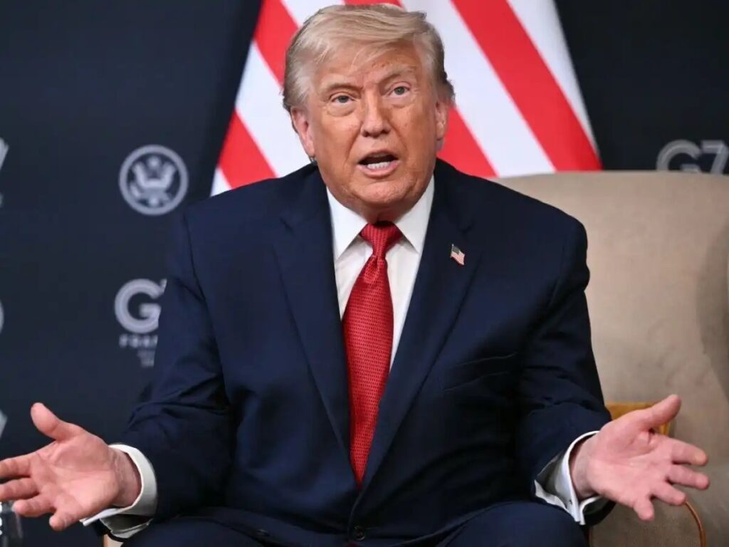 Donald Trump em pronunciamento, sentado em uma cadeira, com as mãos abertas em gesto de fala. Ao fundo, bandeira dos EUA e logotipos em cenário oficial.