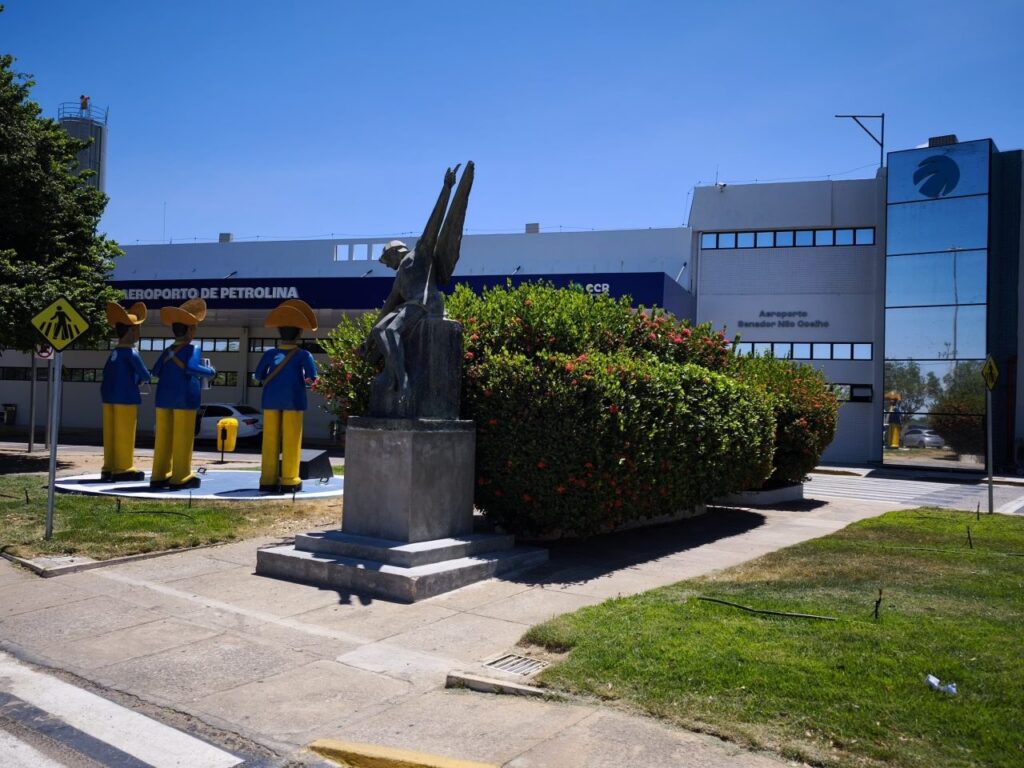 Fachada de um prédio industrial em área externa com jardim, calçada e faixa de pedestres. Em frente, estátuas e funcionários uniformizados próximos a um monumento de pedra sob céu azul.