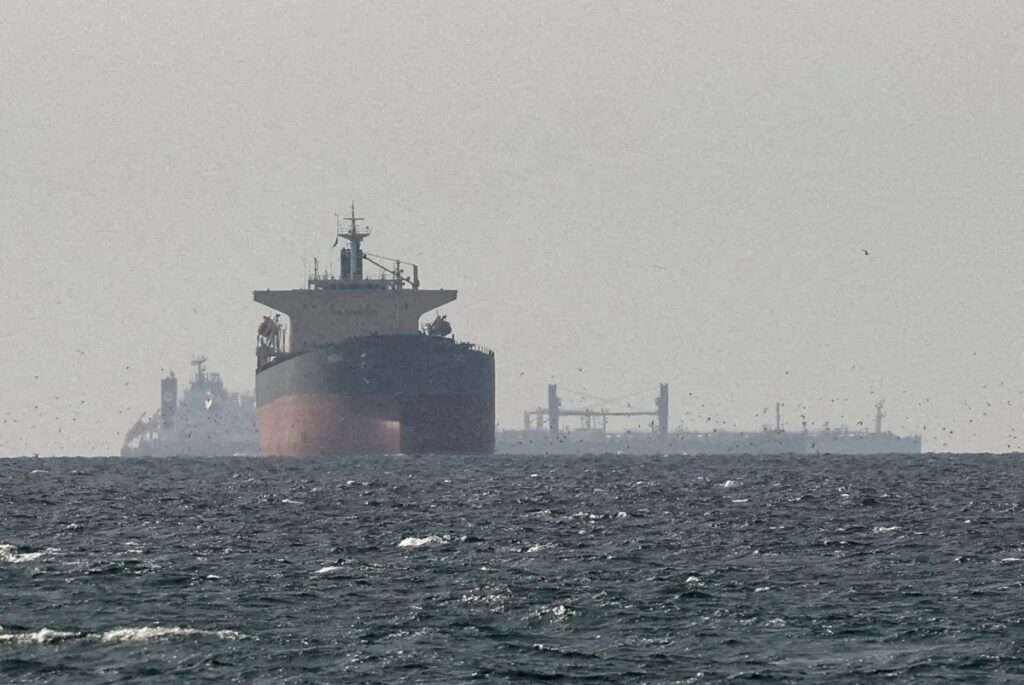 Navio de carga em alto mar, com o casco escuro visível ao fundo e a proa direcionada para a câmera, em cena marítima com ondas e céu nublado