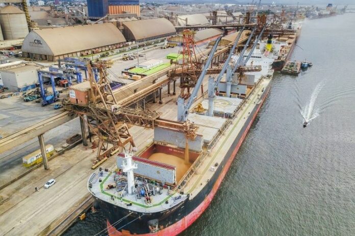 Visão aérea de um navio-tanque em um porto industrial, com guindastes, esteiras e estruturas de carga ao lado da água, mostrando operações logísticas no cais.