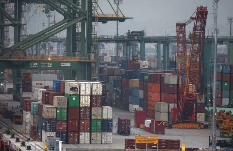 Pátio de contêineres em um porto com grandes guindastes, blocos de carga coloridos e empilhamento de mercadorias sob estrutura industrial, com caminhão e equipamentos ao fundo.
