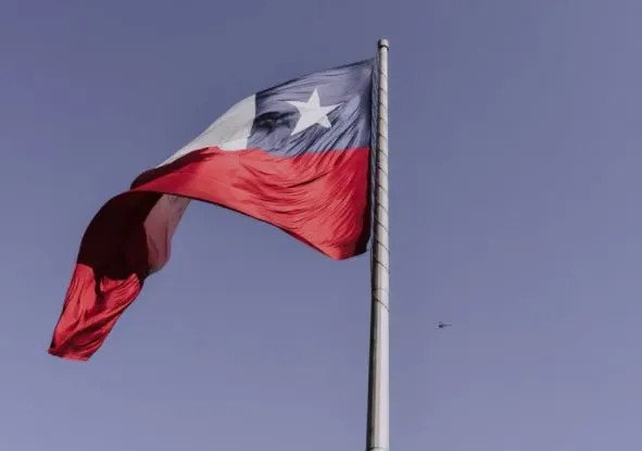 Bandeira do Chile tremulando no alto em um mastro, com cores azul, branca e vermelha, formando o desenho de uma estrela branca ao centro contra o céu claro.
