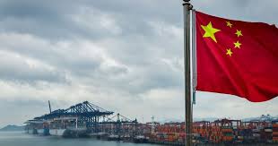 Bandeira da China ao vento em primeiro plano, com um porto industrial ao fundo e guindastes sobre o cais, sob céu nublado