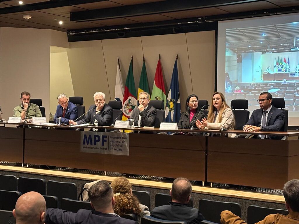Reunião em plenário com autoridades sentadas à mesa, microfones e bandeiras ao fundo, durante sessão de órgão público com telão exibindo pauta em andamento.