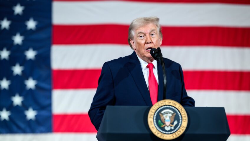Donald Trump discursa em um púlpito diante da bandeira dos Estados Unidos, segurando um microfone e usando terno azul e gravata vermelha em evento político.