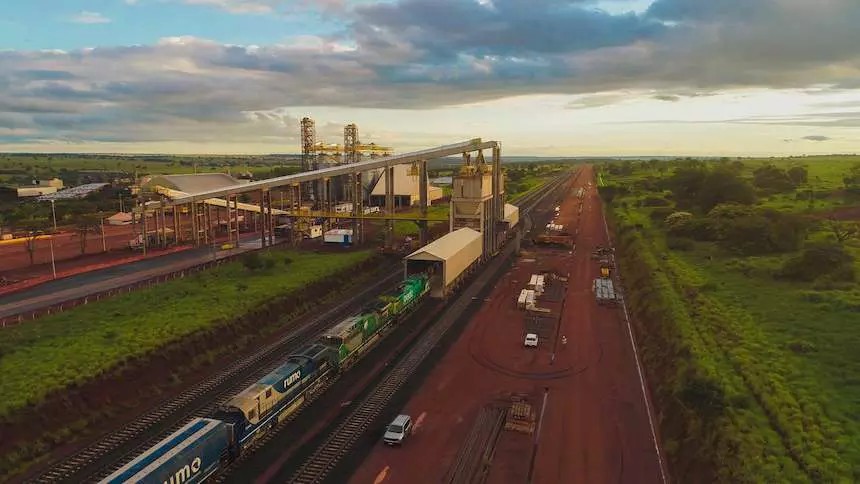 Vista aérea de um terminal ferroviário em operação, com trilhos, pátio de cargas e caminhões em uma estrada de acesso, ao pôr do sol com céu parcialmente nublado e áreas verdes ao redor.