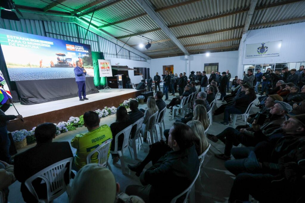 Homem em pé sobre um palco apresenta em um evento corporativo ou institucional em um galpão, com telões e público sentado em cadeiras plásticas, muitos de uniforme, iluminação verde e banners ao fundo.