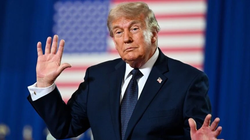 Donald Trump, ex-presidente dos Estados Unidos, aparece em um discurso ou evento, com a bandeira dos EUA ao fundo, levantando a mão em saudação.