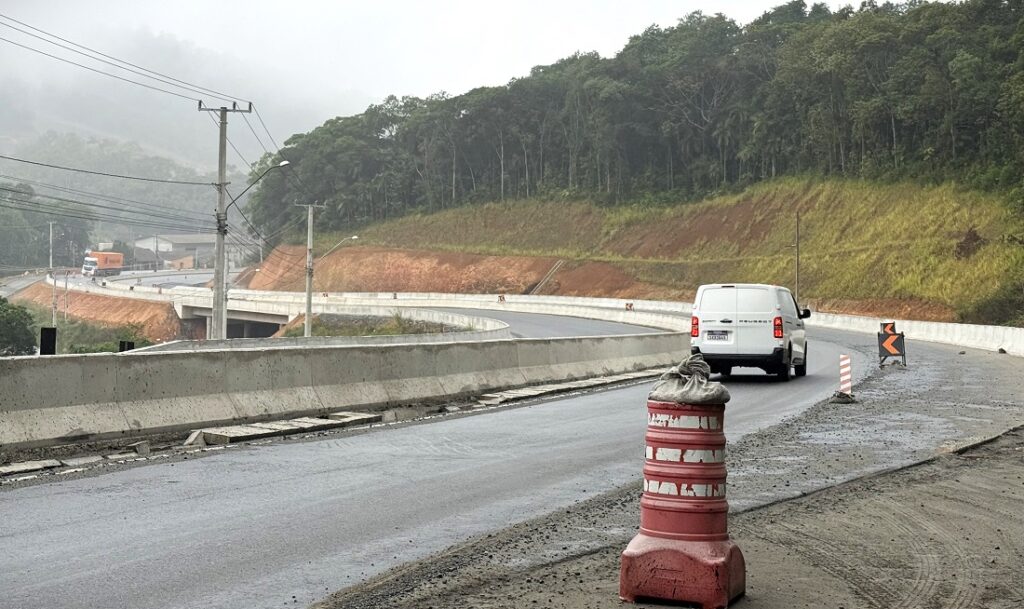 Rodovia em obra com piso molhado e barreira de concreto, cone de sinalização e caminhão branco em curva ao lado de encosta coberta de vegetação.