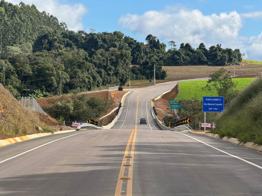 Rodovia em obras com pista recém-asfaltada, faixa central amarela e sinalização de “Ponte sobre o Rio Ribeiro Caçador” indicando extensão de 1,5 km, em meio a mata e colinas.