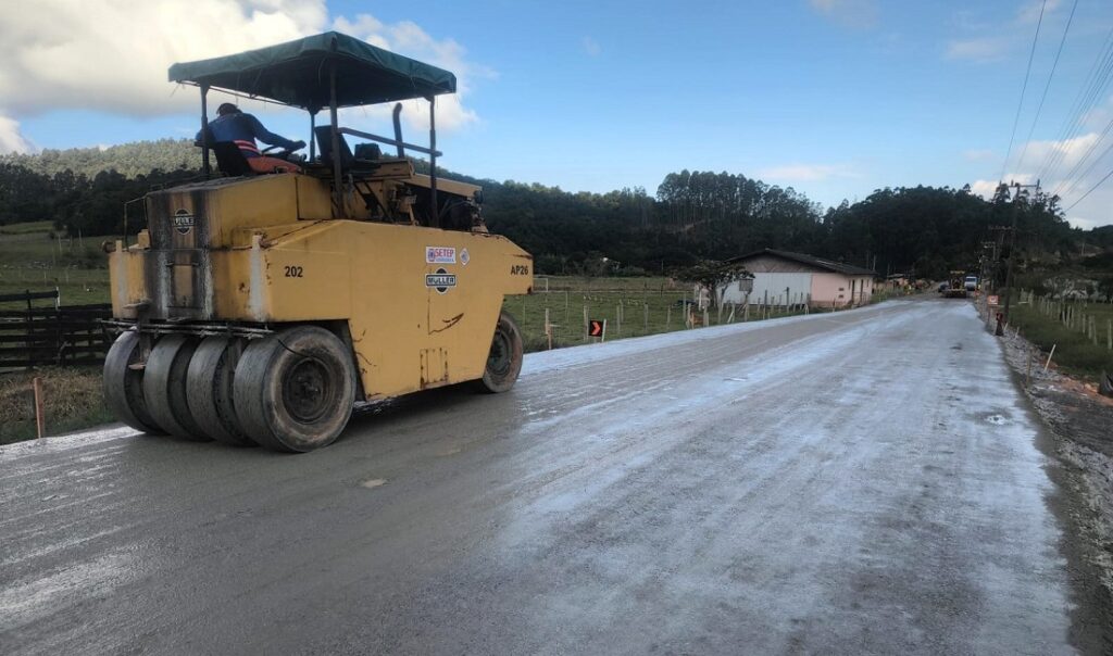 Rolo compactador amarelo asfaltando uma via rural, com operador ao lado da cabine, em uma estrada de concreto asfáltico próxima a cercas e casas, sob céu parcialmente nublado.