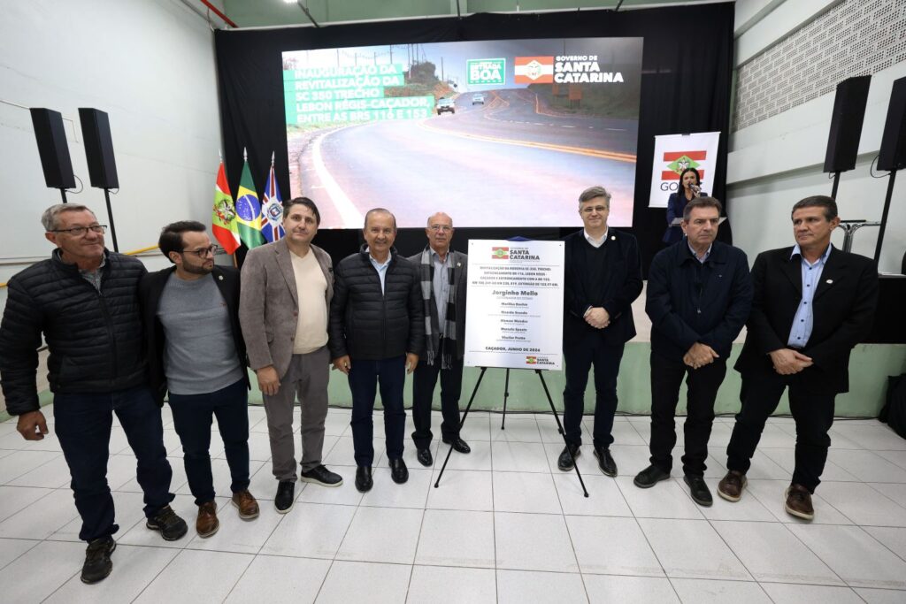 Grupo de autoridades e representantes posa em frente a um telão com informações do Governo de Santa Catarina e um banner na parede, durante evento de lançamento ou apresentação. Ao centro, um painel em suporte com texto e logotipos.