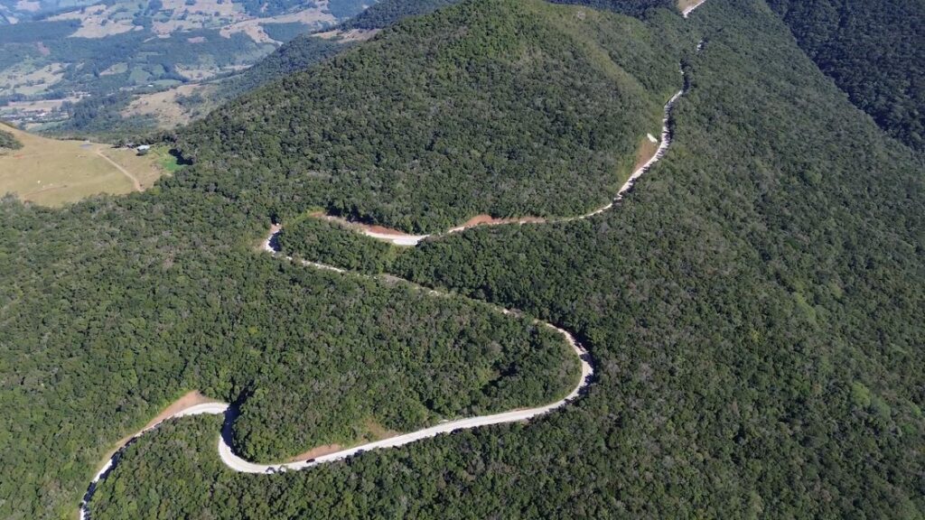 Vista aérea de uma estrada de terra sinuosa atravessando uma grande área de floresta verde e montanhas, com rio e trilhas ao fundo no vale