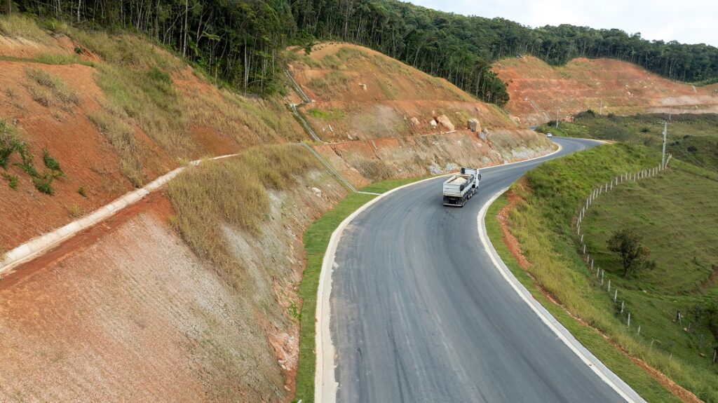 Caminhão trafegando em uma rodovia de asfalto sinuosa entre cortes de terra avermelhada e vegetação de encosta, com cerca ao lado e colinas ao fundo sob céu nublado.