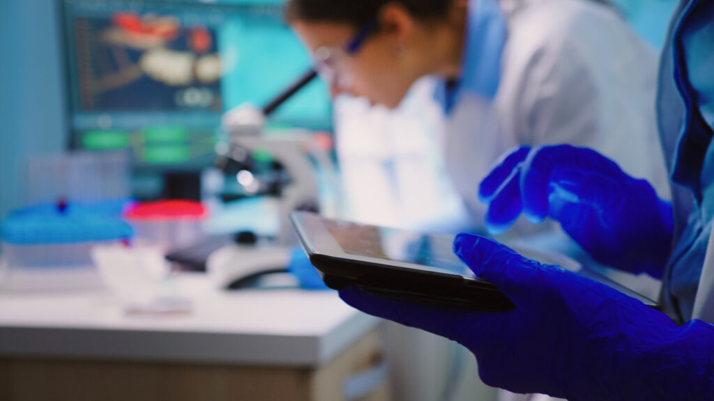 Técnico de laboratório usando luvas azuis e um tablet para analisar resultados em equipamentos de bancada, com monitores e instrumentos ao fundo. Ambiente de pesquisa e controle de qualidade.