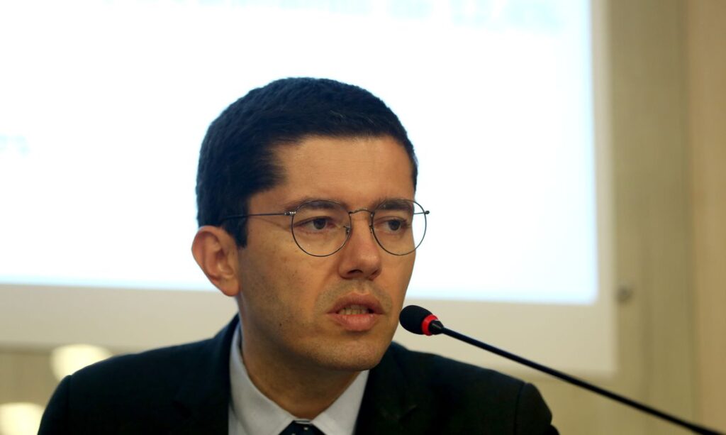 Homem usando óculos de armação metálica e terno escuro em entrevista coletiva, falando ao microfone em uma sala de conferências.