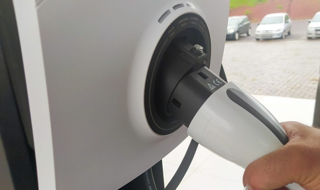 Close-up de um carregador de veículo elétrico conectado a uma tomada de recarga em carro branco, com a mão segurando o plugue durante o processo de carregamento.