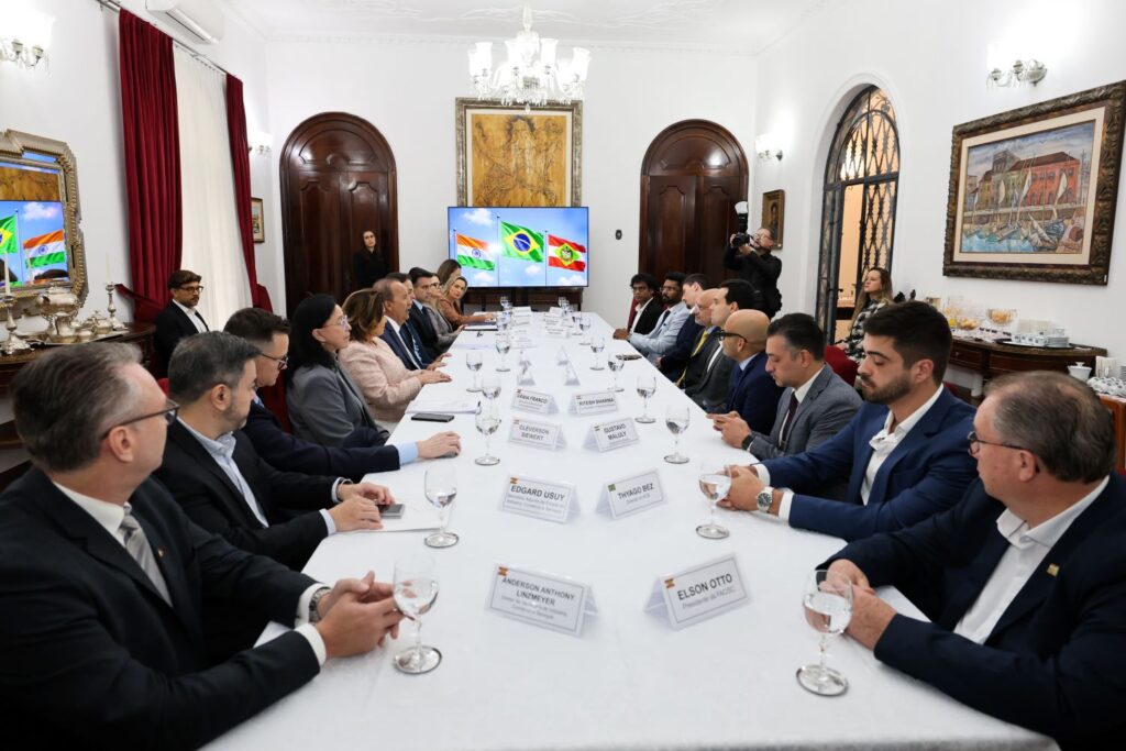 Reunião em sala formal com mesa grande e participantes com taças de vidro, durante encontro diplomático, ao fundo TV exibindo bandeira do Brasil e decoração clássica.