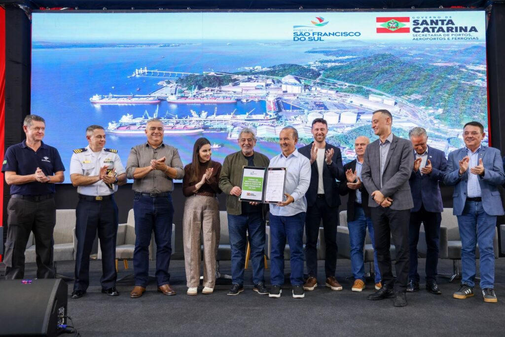 Equipe de autoridades e executivos em evento de assinatura de documentos diante de um telão com imagens do Porto de São Francisco do Sul (Santa Catarina), ao lado de um navio e estruturas portuárias.