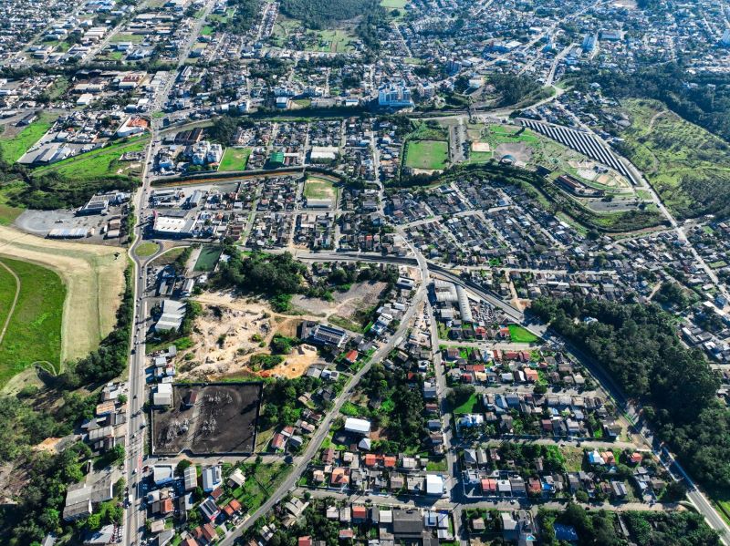 Foto aérea de uma cidade com bairros residenciais, ruas e avenidas, áreas verdes e terrenos baldios, vista do alto com rios e colinas ao fundo.