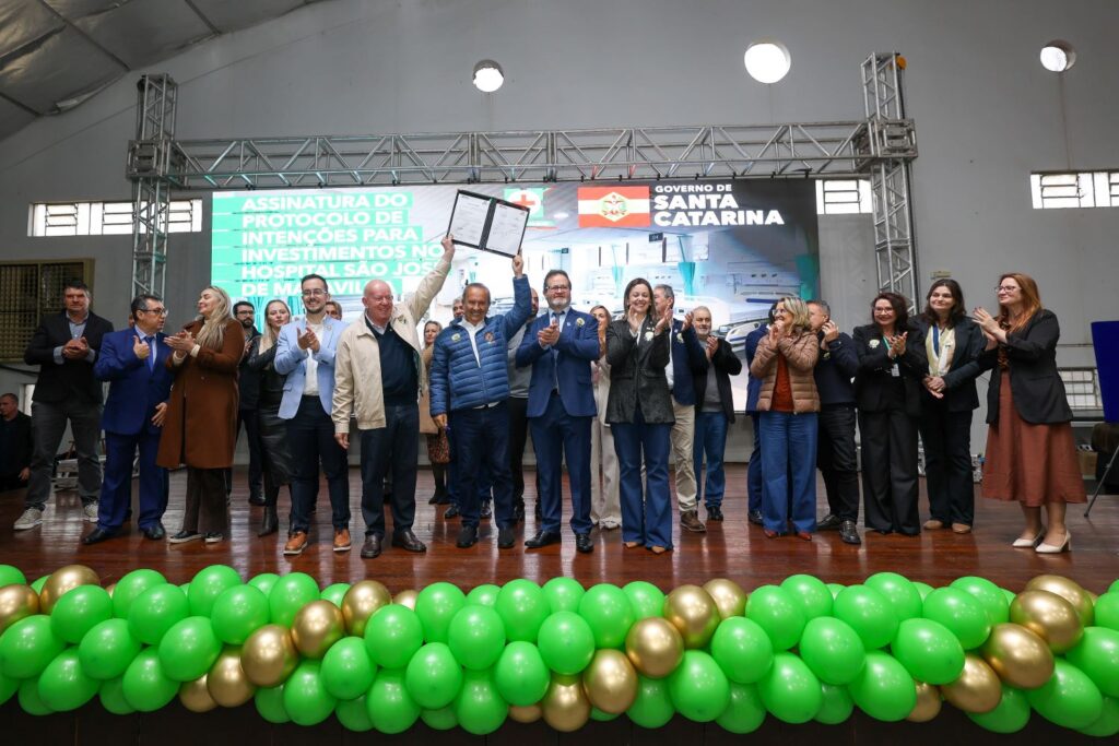 Grupo de autoridades e convidados em evento oficial em Santa Catarina, com assinatura de contrato ao fundo e arco de balões verdes e dourados na frente do palco.