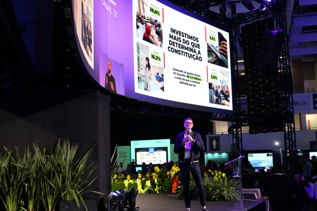 Homem palestrando em palco de evento corporativo, com telões ao fundo mostrando gráficos e texto “Investimos mais do que determina a Constituição”, no auditório iluminado por luzes roxas e verdes.