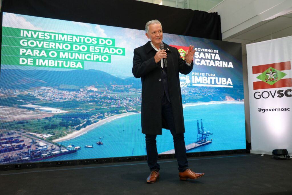 Homem em pé segurando um microfone em evento com telão ao fundo mostrando “Investimentos do Governo do Estado para o município de Imbituba”, cenário com bandeiras e logotipos do Governo de Santa Catarina e Prefeitura de Imbituba.