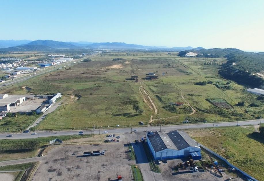 Vista aérea de uma área rural com galpões e instalações industriais ao lado de uma rodovia, cercados por pastagens verdes e montanhas ao fundo, ao entardecer.