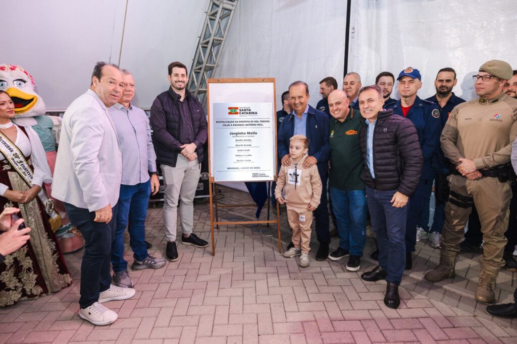 Foto em evento ao ar livre com um painel e placa de inauguração em frente a uma estrutura branca, com autoridades e participantes posando em grupo.