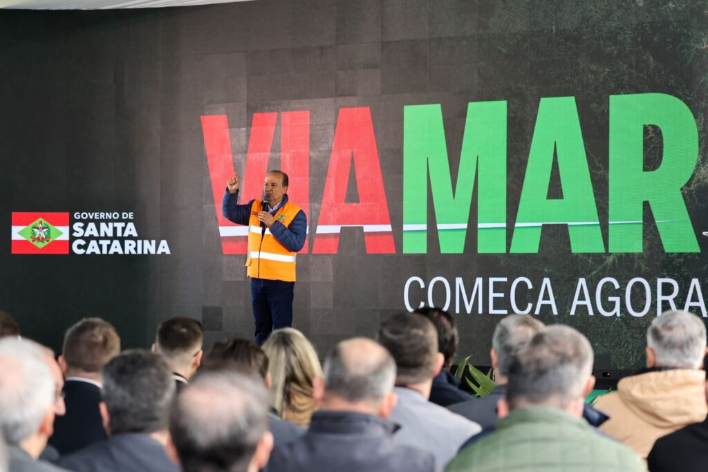 Homem discursando em palco durante evento com plateia, segurando um microfone. Ao fundo há mensagem “WARMAR” e “COMEÇA AGORA”, além do logo do Governo de Santa Catarina.