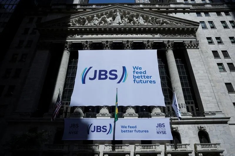 Fachada do prédio da New York Stock Exchange (NYSE) com painel e faixas da JBS, incluindo o texto “We feed better futures” e “JBS listed NYSE”, com bandeira hasteada em frente.