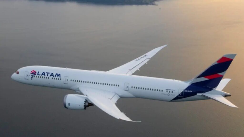Avião da LATAM em voo no céu, com pintura branca e detalhes azul e vermelho nas asas, fotografado em perspectiva lateral mostrando as turbinas e as janelas da cabine.
