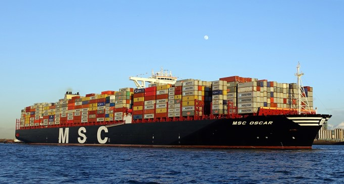 Navio porta-contêiner MSC Oscar em navegação no mar, com grande quantidade de contêineres empilhados no convés e letras MSC na lateral sob céu azul claro.