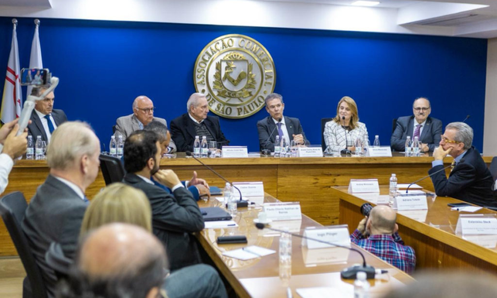Reunião em plenário com pessoas sentadas ao redor de uma mesa, durante sessão na sede da Associação Comercial de São Paulo (ACSP), com membros ao fundo diante do brasão.