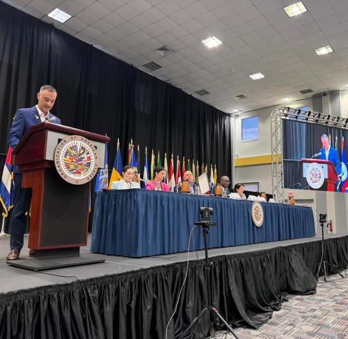Homem discursando em um púlpito de madeira em uma sala de conferências, com bandeiras ao fundo e uma mesa de painel ao lado, em evento institucional