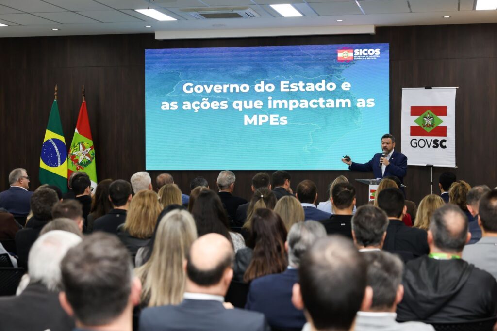 Homem palestrando em auditório lotado, ao lado de banner com a mensagem “Governo do Estado e as ações que impactam as MPEs”, com bandeiras do Brasil e de Santa Catarina e logomarca SICOS no telão.