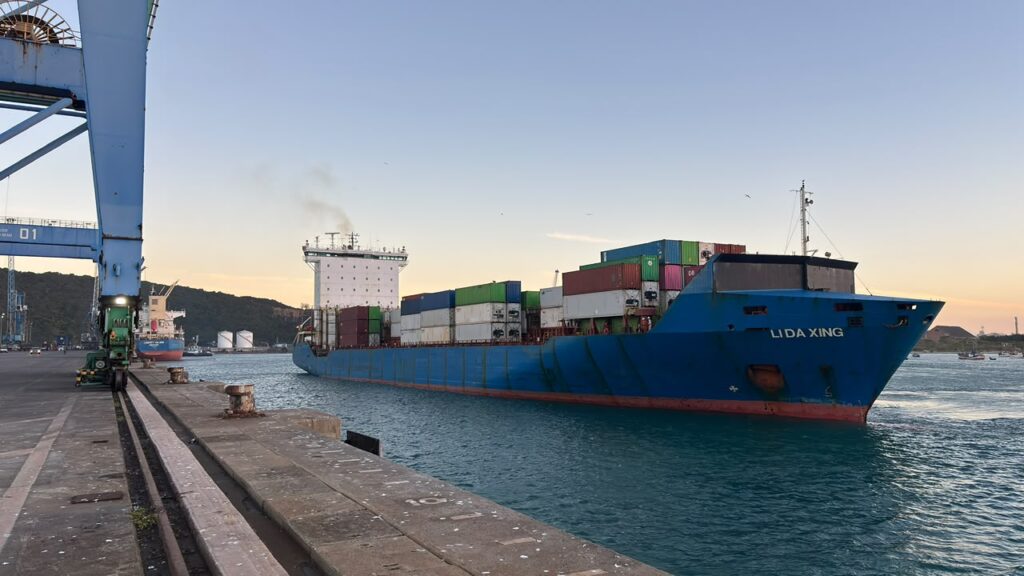 Navio de carga azul ancorado em um porto ao entardecer, com contêineres coloridos no convés e estruturas portuárias ao lado do cais, em cenário de mar calmo