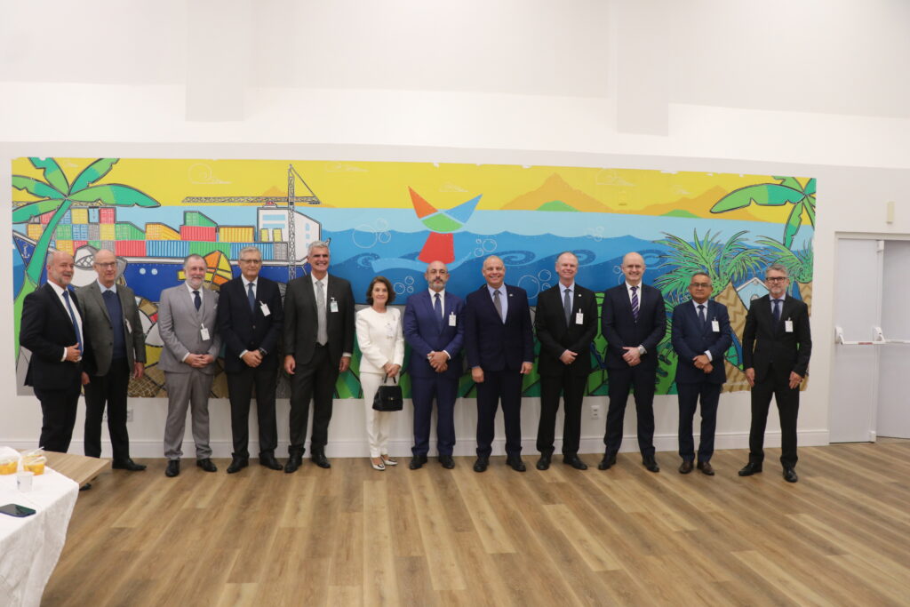 Grupo de pessoas em reunião oficial com traje social posando em frente a um mural artístico de paisagem com mar, palmeiras e prédios coloridos em um ambiente interno