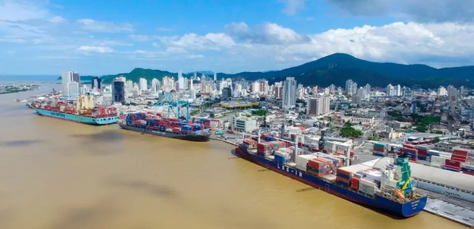 Vista aérea de um porto com navios cargueiros atracados ao longo do cais e contêineres, com skyline urbano ao fundo e montanhas na paisagem, em um dia nublado.