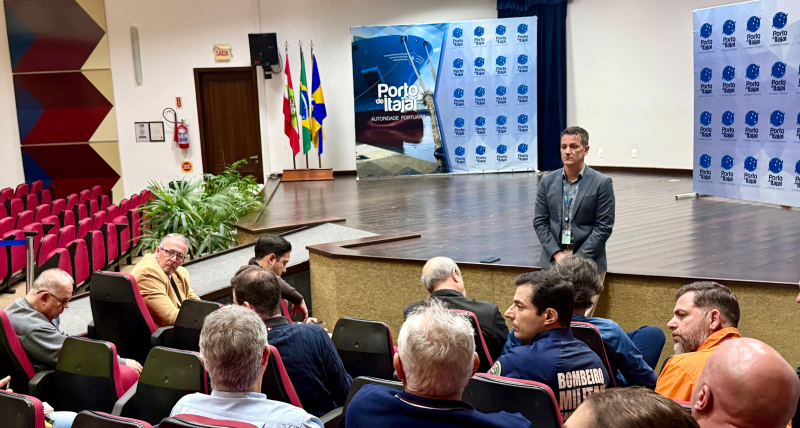 Palestra em auditório com plateia sentada e banner “Porto de Itajaí” ao fundo, com palestrante em pé em um evento institucional. Bandeiras e telões reforçam o contexto do encontro.