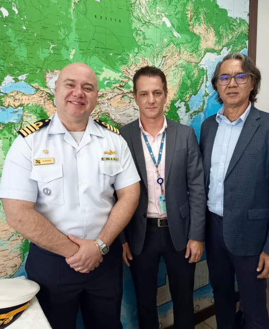 Três pessoas posam para foto em evento, diante de um mapa-múndi, com um militar uniformizado à esquerda, um homem de terno no centro e uma mulher de blazer à direita.