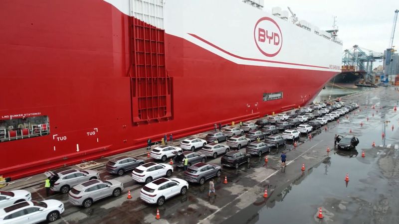 Navio porta-veículos com a marca BYD transportando carros alinhados no convés, em operação no porto, com equipe e cones de sinalização na área de carregamento