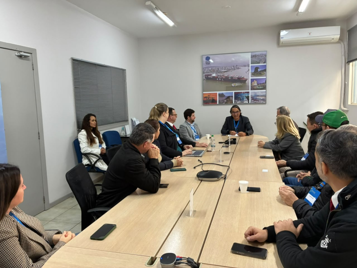 Reunião em sala de treinamento com participantes sentados em volta de uma mesa, notebooks e celulares sobre a superfície, ao lado de um painel com fotos na parede e ar-condicionado no teto.