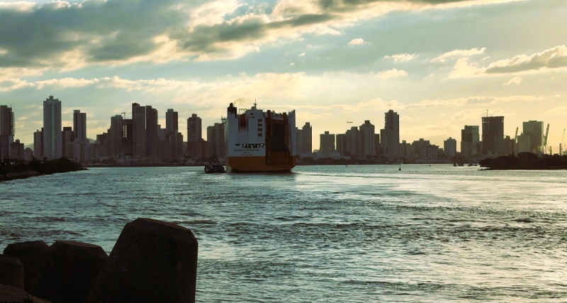 Vista do skyline urbano ao pôr do sol com um navio cargueiro ancorado no rio, ondas refletindo a luz dourada e prédios altos ao fundo sob céu parcialmente nublado.