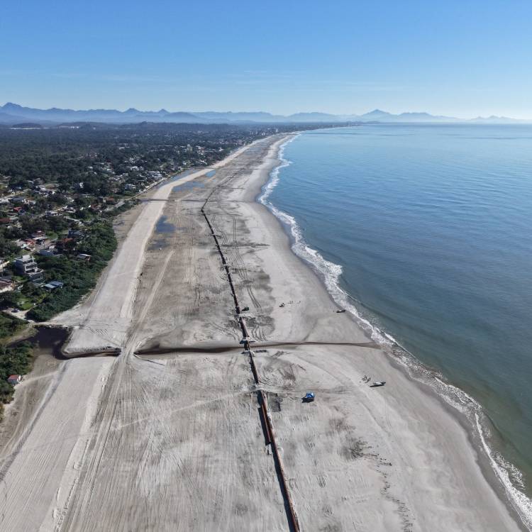 Vista aérea de um trecho de rodovia e canteiro de obras na costa, com praia de areia ao lado do mar calmo, nuvens baixas e montanhas ao fundo.