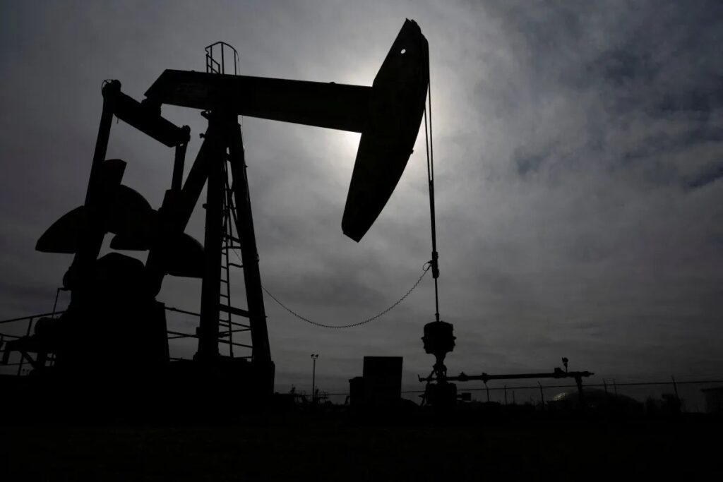 Silhueta de um poço de petróleo com uma bomba mecânica em funcionamento, vista contra o céu nublado ao entardecer, com tubulação e estruturas industriais ao fundo.